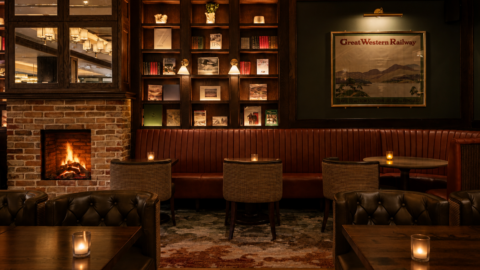 Dimly lit lounge with leather chairs, small tables, a brick fireplace, bookshelves, and a &ldquo;Great Western Railway&rdquo; wall art. Several candles are placed on the tables, creating a cozy atmosphere.