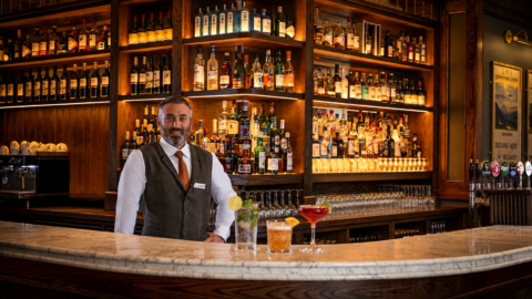 A bartender in a vest stands behind a marble bar counter with three drinks; shelves filled with various liquor bottles are in the background.