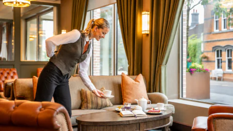 A server places a teacup on a table with a teapot, a notebook, and snacks in a cozy, well-lit lounge with large windows and plush seating.