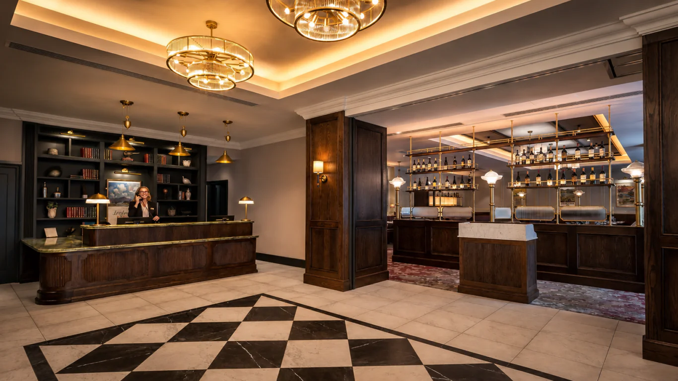 Hotel lobby with a reception desk on the left, a staff member on the phone, and a bar area with shelves of bottles on the right. The room features warm lighting and geometric tile flooring.