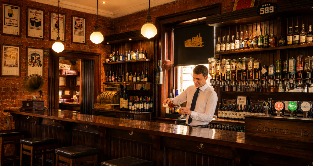 signal bar 1 warm A bartender in a white shirt and tie pours a drink behind a wooden bar in a cozy, warmly lit pub with bottles and framed pictures on the brick walls.