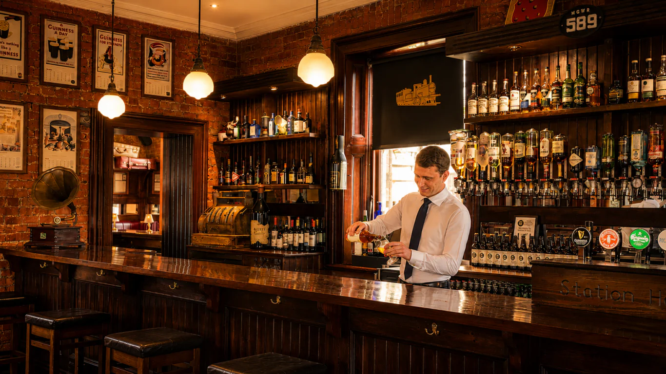 A bartender in a white shirt and tie pours a drink behind a wooden bar in a cozy, warmly lit pub with bottles and framed pictures on the brick walls.