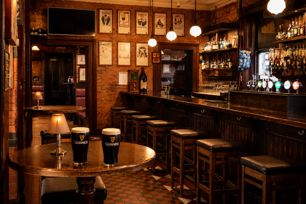 signal bar warm Dimly lit pub interior with a wooden bar, high stools, framed pictures on brick walls, and two pints of Guinness on a round table in the foreground.