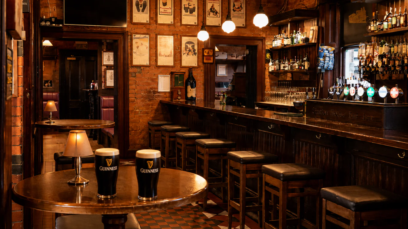 Dimly lit pub interior with a wooden bar, high stools, framed pictures on brick walls, and two pints of Guinness on a round table in the foreground.