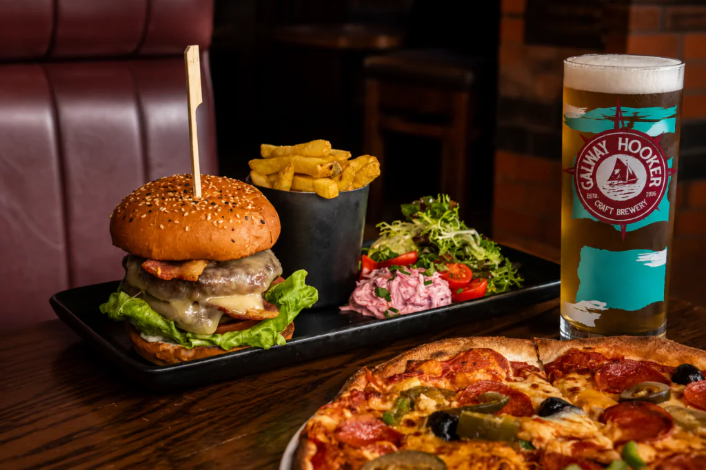 signal burger and pizza A cheeseburger with lettuce, fries, salad, a glass of beer, and part of a pizza on a wooden table in a restaurant setting.