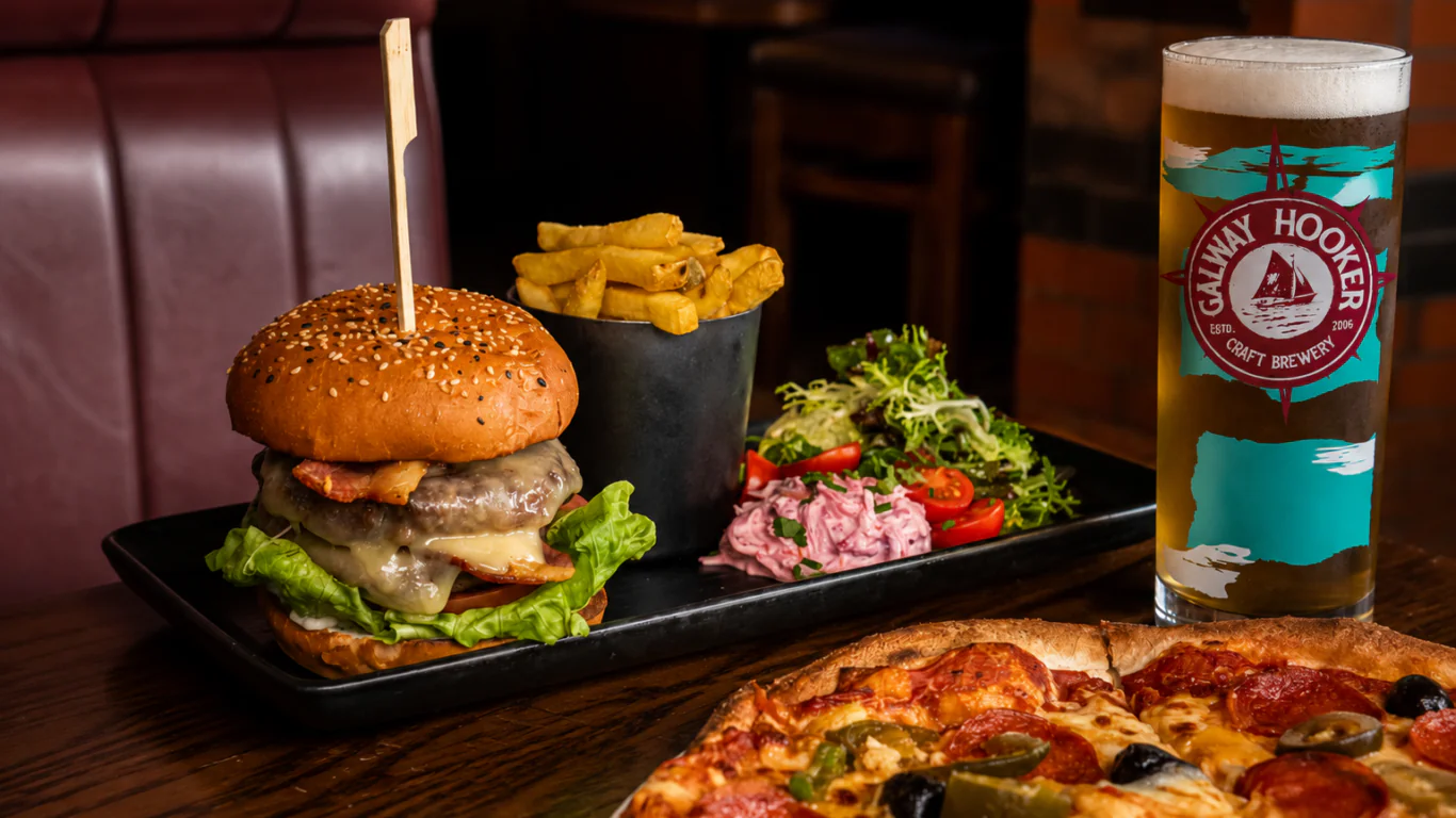 A cheeseburger with lettuce, fries, salad, a glass of beer, and part of a pizza on a wooden table in a restaurant setting.